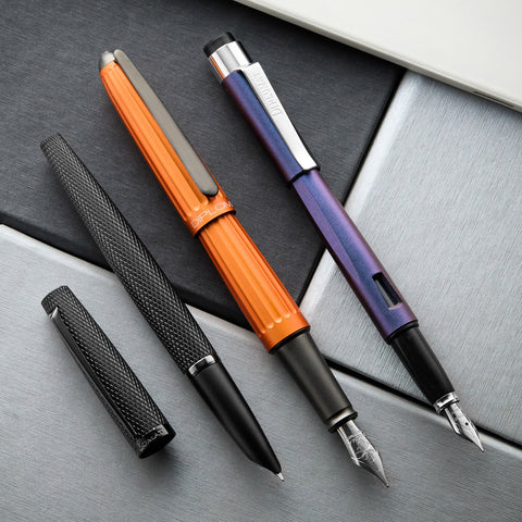 Close up of several fountain pens on a dark desk, including a bright orange Diplomat pen.