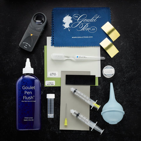 Goulet Pens product shot with a blue microfiber cloth, Goulet Pen Flush bottle, and assorted pen cleaning tools on a dark surface