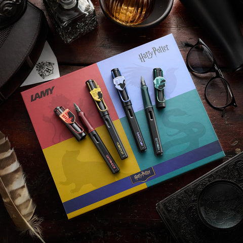 Lamy x Harry Potter fountain pens arranged on color block notebooks with a feather quill and reading glasses on a dark wooden desk