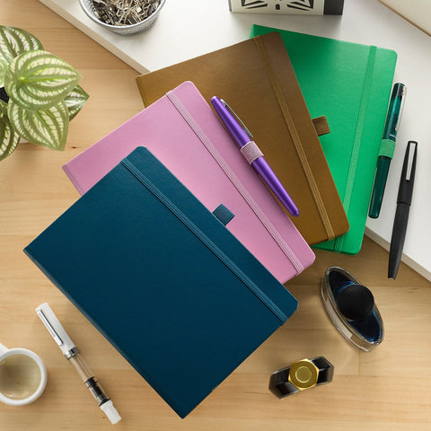 Leuchtturm1917 notebooks in teal navy pink brown and green with a purple pen on top and other office supplies on a wooden desk