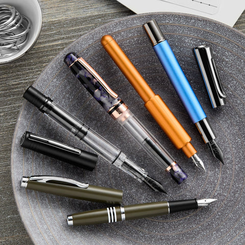 Monteverde fountain pens in orange blue black and marbled purple arranged diagonally on a speckled plate over a wooden surface
