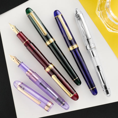 Platinum fountain pens in green black purple and clear with gold trim arranged on a white desk beside a bright yellow notebook