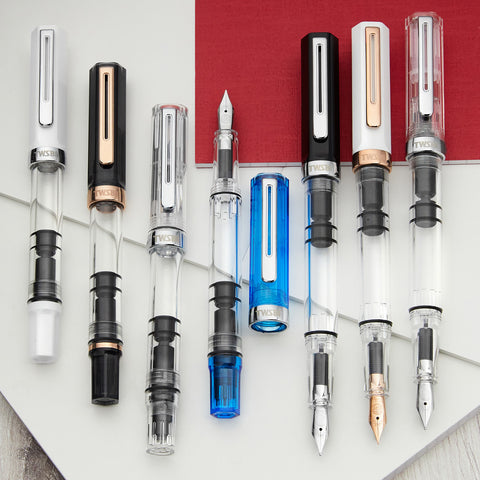 Row of piston fountain pens in white black clear and blue finishes displayed on a white backdrop with a red panel