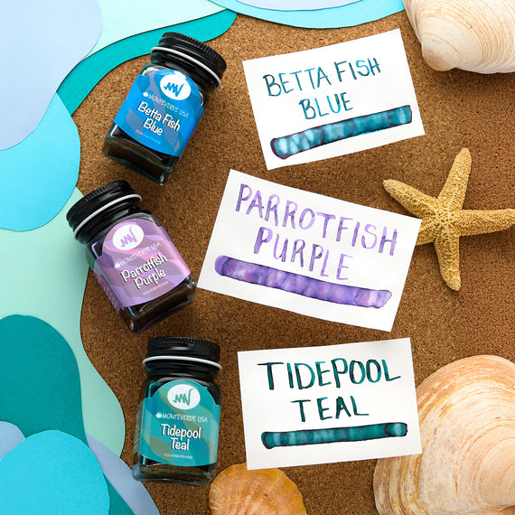 Three ink bottles labeled 'Betta Fish Blue', 'Parrotfish Purple', and 'Tidepool Teal' on a cork board with starfish and shells.