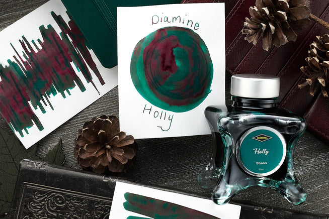 Diamine Holly Fountain Pen Ink writing sample, and swabs, along iwth the bottle and pinecones on a wooden desk background