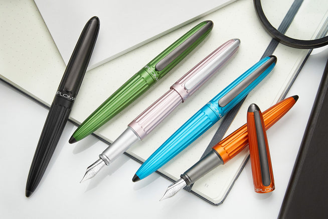 5 Diplomat Aero Fountain Pens in black, green, pink, turquoise, and orange on a notebook/white background. Two pens have the nibs showing.