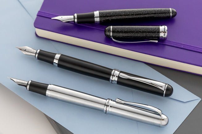 3 uncapped Jinhao fountain pens in silver, black and shiny on blue envelope and purple journal