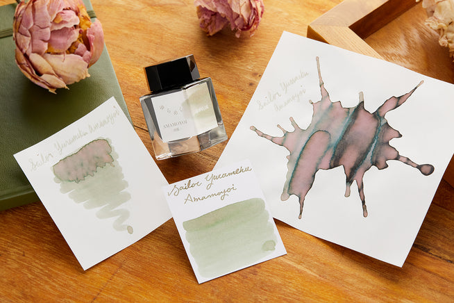Three white pieces of paper with Sailor Yurameku Amamoyoi Fountain Pen ink on each card in a different shape or pattern and glass product bottle in between them.