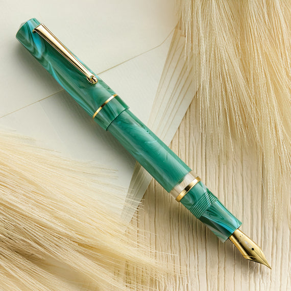 The image shows a fountain pen with a green and gold patterned body and a posted cap. The pen has gold trim and clip, and a conical tip.