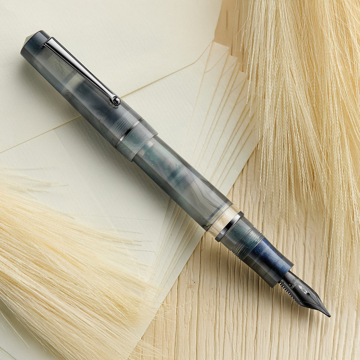 The image shows a Delta Duna Zephyr fountain pen. It has a gray and blue translucent body with a mother-of-pearl finish, ruthenium trim, and a matching nib.