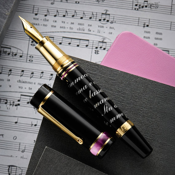 A fountain pen made of hand-turned, mirror-polished resin in black with pink accents, with cursive white writing on the barrel.