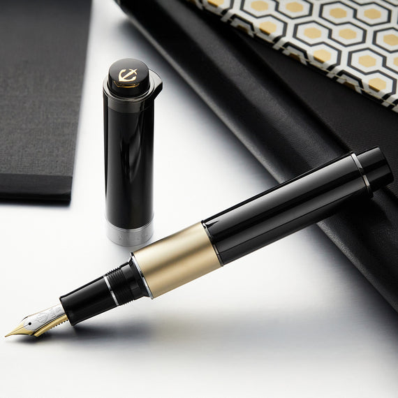 Black and gold fountain pen on a reflective surface with a geometric-patterned object in the background