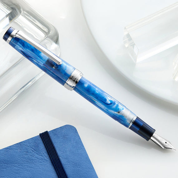 Blue fountain pen on a white surface with a clear glass object in the background